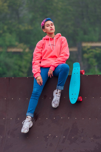 Portrait of a trendy pretty young girl with short colored hair and nose piercing dressed in pink hoodie and jeans sitting next to the skateboard court with her blue plastic skateboard. - Фото, изображение