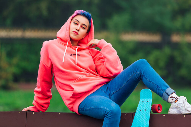 Portrait of a trendy pretty young girl with short colored hair and nose piercing dressed in pink hoodie and jeans sitting next to the skateboard court with her blue plastic skateboard. - Fotografie, Obrázek
