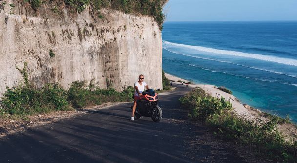 Un joven con gafas se sienta en una motocicleta deportiva roja contra el fondo del mar - Foto, Imagen