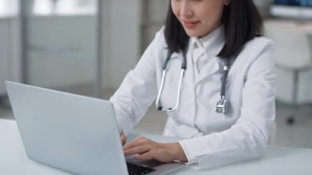 Portrait of beautiful young adult Asian woman wearing white coat and stethoscope around her neck working on laptop then smiling at camera - Footage, Video