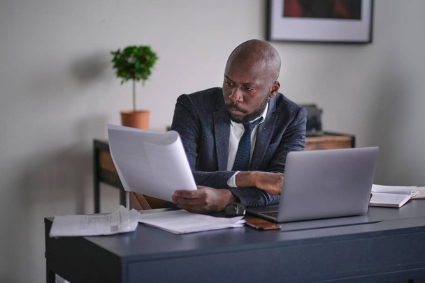 A fiatal afrikai vezető dokumentumokat olvas és laptopot javít, miközben az íróasztalánál ül egy irodában. lágy fókusz - Fotó, kép