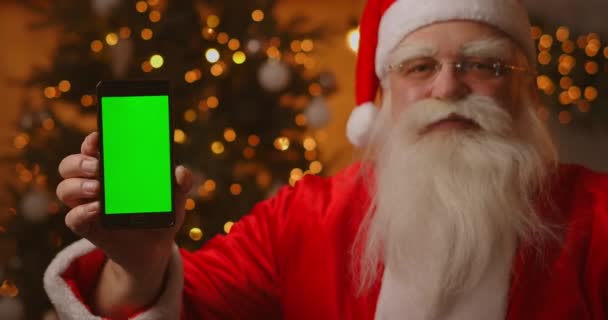Święty Mikołaj siedzi na kanapie w tle choinki i girlandy trzymając telefon komórkowy z zielonym ekranem wskazując na nią palcem. - Materiał filmowy, wideo