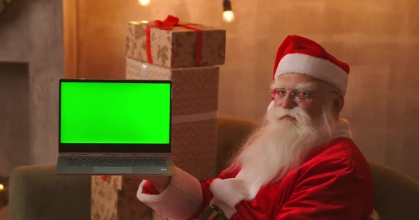 Mikulás tart egy laptop zöld képernyővel, miközben ül a karácsonyi dekorációk. - Felvétel, videó
