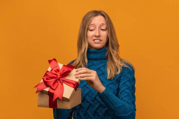 Charmante jeune femme blonde en pull bleu tricoté ouvrir un cadeau avec ruban rouge. Plan studio, fond jaune. Nouvel An, Fête des femmes, Anniversaire, Concept vacances - Photo, image