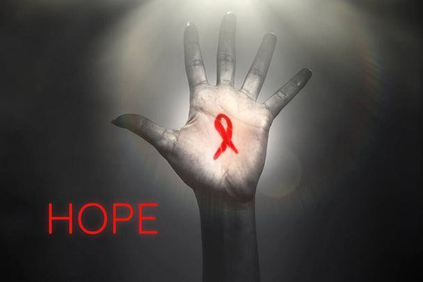 Το χέρι μιας γυναίκας φτάνει στο φως με μια κόκκινη κορδέλα στην παλάμη. Σκούρο φόντο με φως στην κορυφή. Η έννοια της παγκόσμιας ημέρας καταπολέμησης του AIDS και η ελπίδα για θεραπεία. - Φωτογραφία, εικόνα