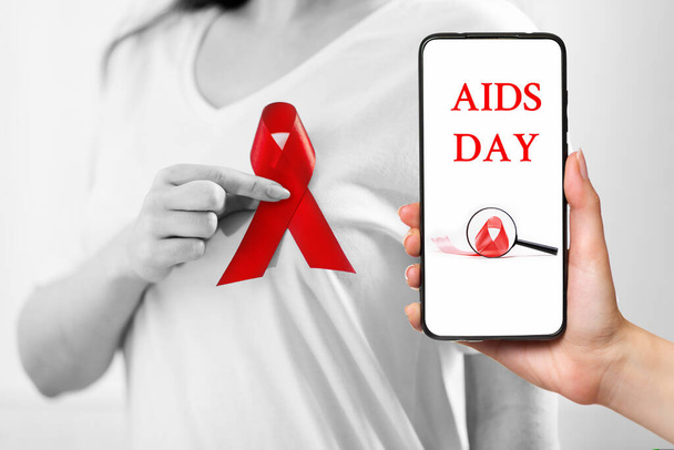 Una mujer sostiene una cinta roja en el pecho con los dedos. Manos de cerca. La mano derecha sostiene un smartphone. El concepto del día mundial del sida. - Foto, imagen