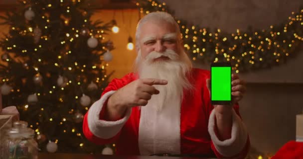 Der Weihnachtsmann sitzt auf dem Sofa im Hintergrund eines Weihnachtsbaums und hält ein Handy in der Hand, auf das ein grüner Bildschirm mit dem Finger zeigt.. - Filmmaterial, Video