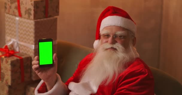 Papai Noel está sentado no sofá no fundo de uma árvore de Natal e guirlandas segurando um telefone celular com uma tela verde apontando para ele com o dedo. - Filmagem, Vídeo
