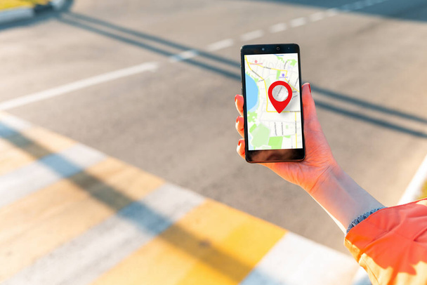 De hand van een vrouw houdt een smartphone vast met een online kaart met een rood geolocatiepictogram. Op de achtergrond, een voetgangersoversteek in een waas. Begrepen, ruimte. Concept van online navigatie en GPS. - Foto, afbeelding