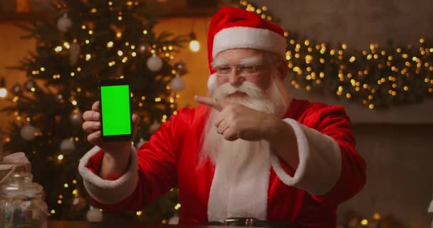 Papai Noel está sentado no sofá no fundo de uma árvore de Natal e guirlandas segurando um telefone celular com uma tela verde apontando para ele com o dedo. - Filmagem, Vídeo
