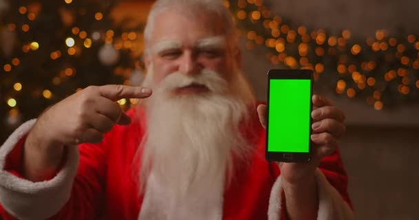 Joulupukki istuu sohvalla joulukuusen ja seppeleiden taustalla pitäen matkapuhelinta, jossa on vihreä näyttö, joka osoittaa sitä sormellaan.. - Materiaali, video