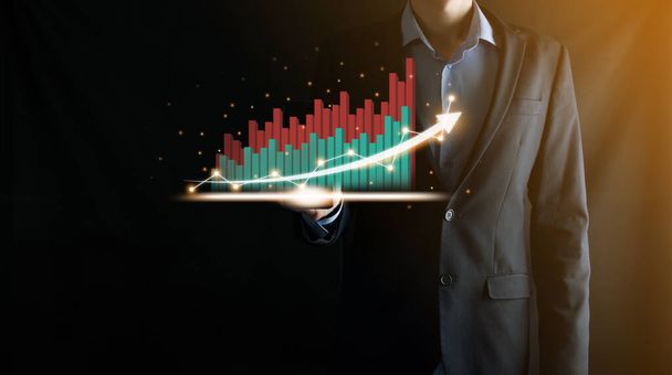 Businessman holding and showing a growing virtual hologram of statistics, graph and chart with arrow up on dark background. Stock market. Business growth, planing and strategy concept - Photo, Image
