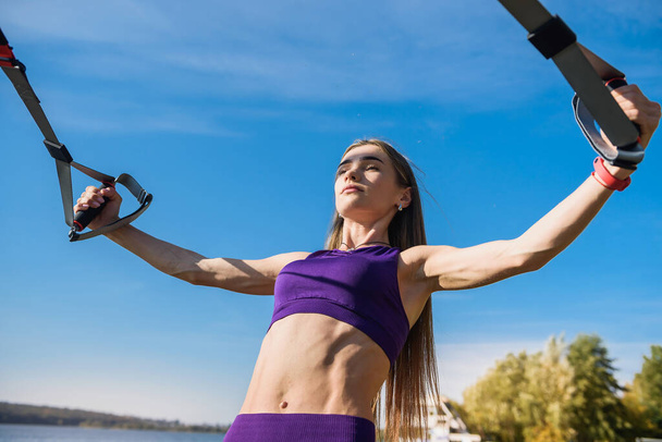 Giovane donna che esercita con sospensione trainer fionda nel parco, vicino al lago. - Foto, immagini