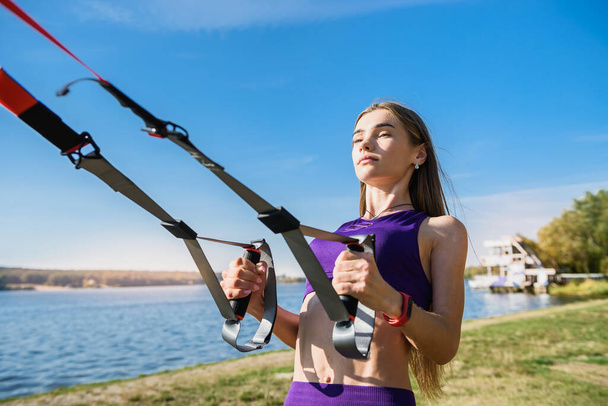 スポーツウェアを身に着けているかなりの女性は、日中に湖の近くに屋外でtrxフィットネスストラップを押し上げています。健康的なライフスタイル - 写真・画像