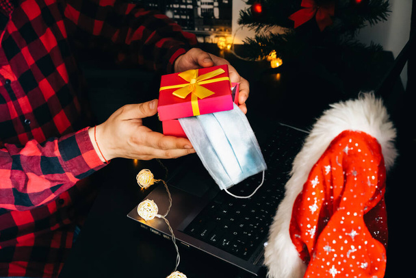 クリスマスプレゼントだ。人はビデオ通話を通して箱のギフトを共有する。隔離中の休日。ウイルスの医療マスク。装飾が施された新年の木とライト。オンラインで祝う家族や友人. - 写真・画像