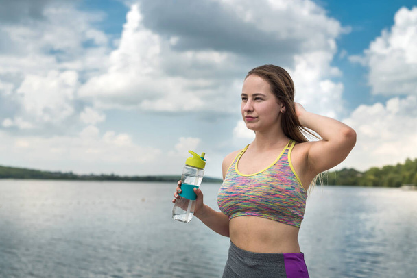 Bella giovane sportiva che riposa vicino al lago dopo l'allenamento con una bottiglia d'acqua in mano. Stile di vita sano - Foto, immagini