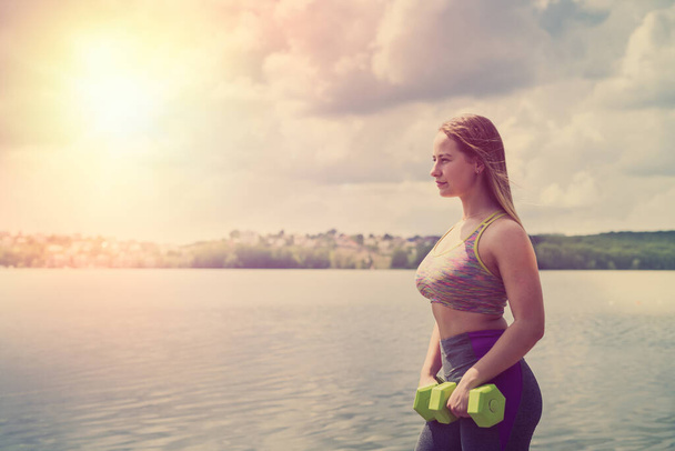 Jovem menina fitness fazendo exercícios com halteres no pôr do sol perto do lago no parque da cidade - Foto, Imagem