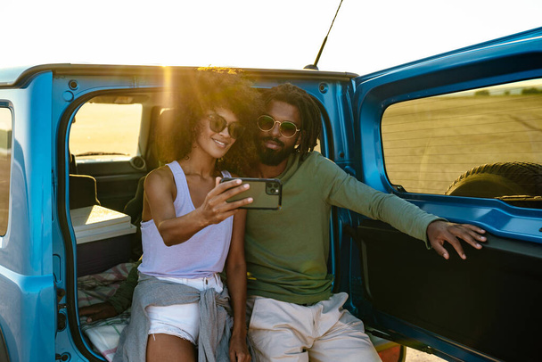 Young couple in love making selfie using smart phone while sitting on a back of car - Photo, image