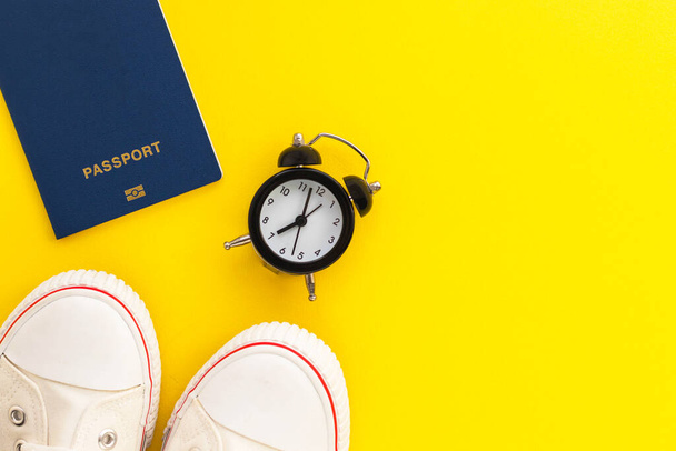 Pasaport, çalar saat ve sarı arka planda sakız ayakkabıları. Seyahat zamanı. Seyahat aksesuarları. Tatil ve tatil konsepti. Üst görünüm, kopyalama alanı olan düz uzanma. - Fotoğraf, Görsel