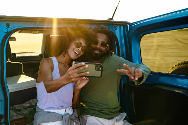 Young couple in love making selfie using smart phone while sitting on a back of car - Fotografie, Obrázek
