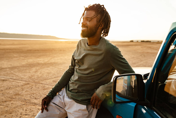 Image d'un homme afro-américain joyeux souriant et voyageant en voiture dans le désert - Photo, image