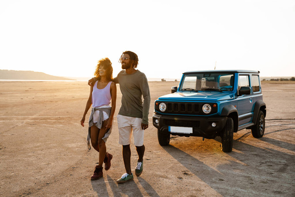 Image of joyful african american couple hugging and walking while travelling with car on desert - Foto, Imagen
