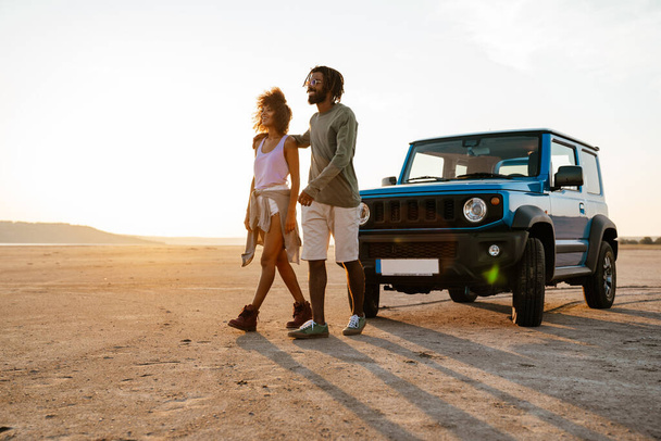 Image of joyful african american couple hugging and walking while travelling with car on desert - Foto, Bild