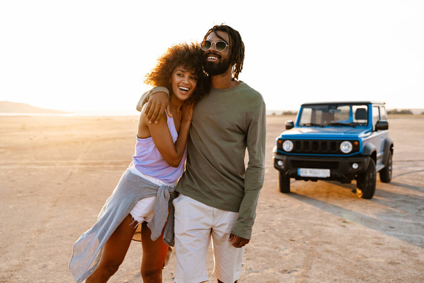 Image of joyful african american couple hugging and walking while travelling with car on desert - Фото, зображення