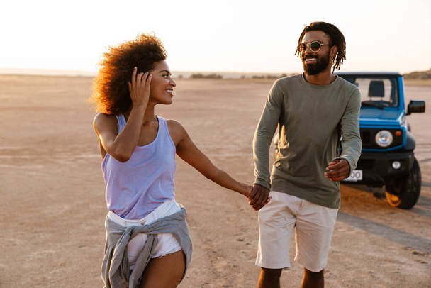 Image of joyful african american couple holding hands together and walking while travelling with car on desert - Photo, image
