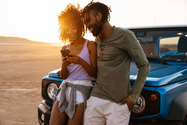 Image of joyful african american couple using mobile phone while travelling with car in desert - Zdjęcie, obraz