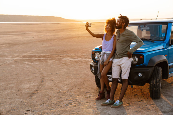 Image of joyful african american couple taking selfie on cellphone while travelling with car in desert - Photo, image