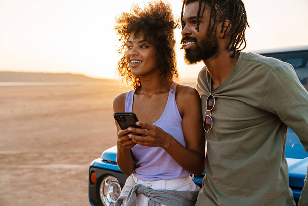 Image of joyful african american couple using mobile phone while travelling with car in desert - Foto, imagen