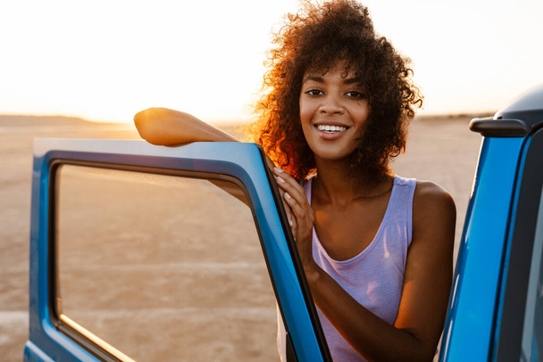 Image of cheerful african american woman smiling while travelling with car on desert - Фото, изображение