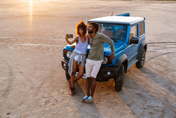 Image of joyful african american couple taking selfie on cellphone while travelling with car in desert - Zdjęcie, obraz