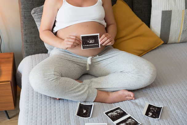 Беременная женщина держит фотографию ультразвука на животе в постели дома. Концепция дородового медицинского обслуживания. - Фото, изображение
