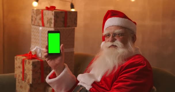Santa Claus is sitting on the sofa in the background of a Christmas tree and garlands holding a mobile phone with a green screen pointing at it with his finger. - Footage, Video