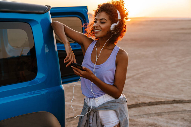 Image of happy african american woman using headphones and cellphone while travelling with car on desert - Foto, afbeelding