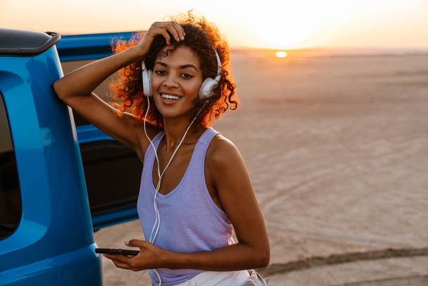 Image of happy african american woman using headphones and cellphone while travelling with car on desert - Fotó, kép