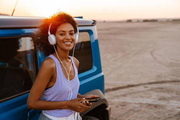 Image of happy african american woman using headphones and cellphone while travelling with car on desert - Photo, Image