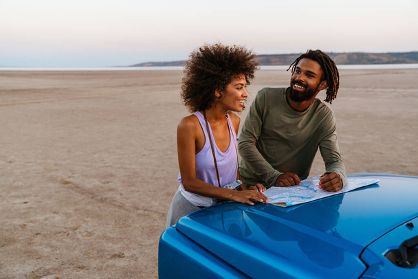 Image of joyful african american couple examining map while travelling with car on desert - Photo, image