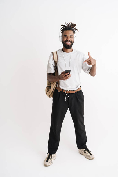 Photo of joyful african american guy in headphones pointing finger at his cellphone isolated over white background - Photo, image