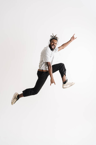 Photo of excited african american guy in headphones jumping while showing horn gesture isolated over white background - Foto, afbeelding