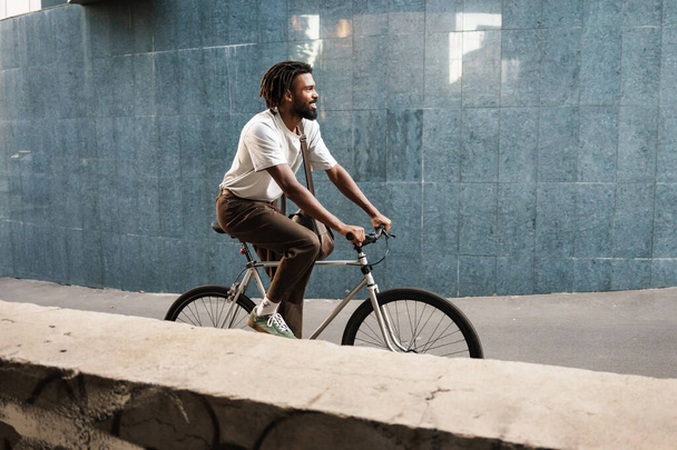 Happy african american guy smiling while riding bicycle at city street - Foto, afbeelding