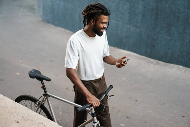 Smiling african american guy using cellphone while walking with bicycle at city street - 写真・画像