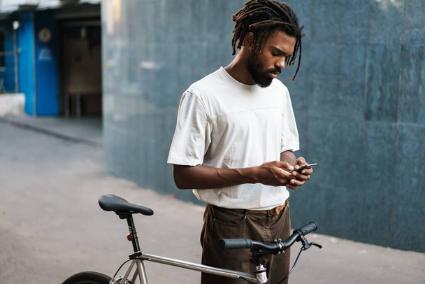 Concentrated african american guy using cellphone while walking with bicycle at city street - Fotoğraf, Görsel