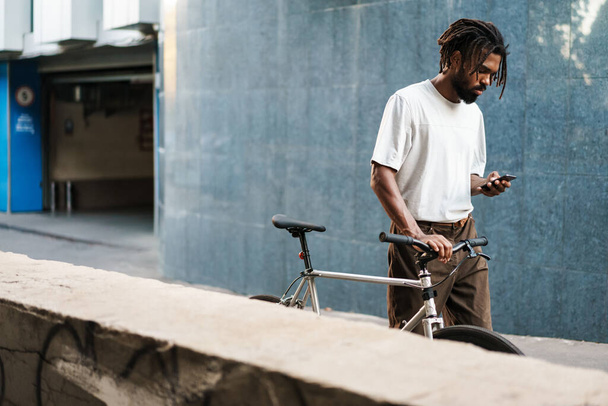 Concentrated african american guy using cellphone while walking with bicycle at city street - 写真・画像