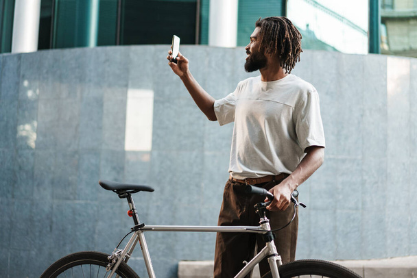 Smiling african american guy taking selfie on cellphone while standing with bicycle at city street - Foto, Imagen