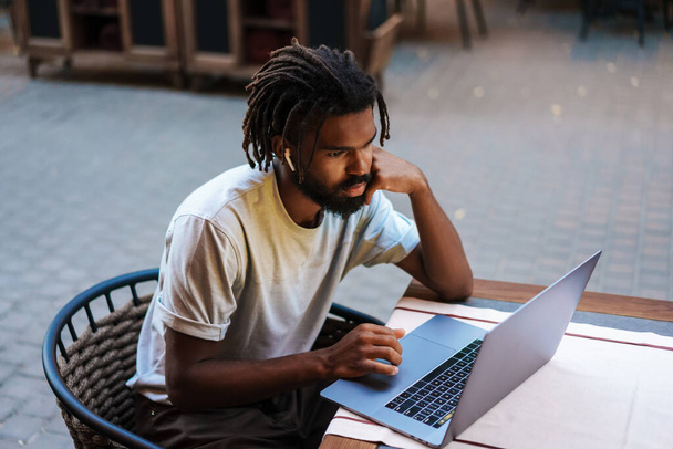Focused african american guy in earphones working with laptop while sitting in cafe outdoors - Фото, изображение