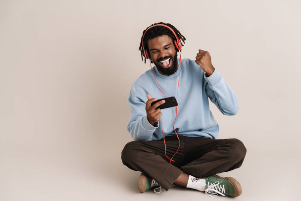 Excited african american guy in headphones using cellphone and making winner gesture isolated over white wall - Valokuva, kuva