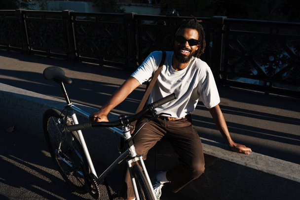 Happy african american guy smiling while posing with bicycle at city street - Fotoğraf, Görsel